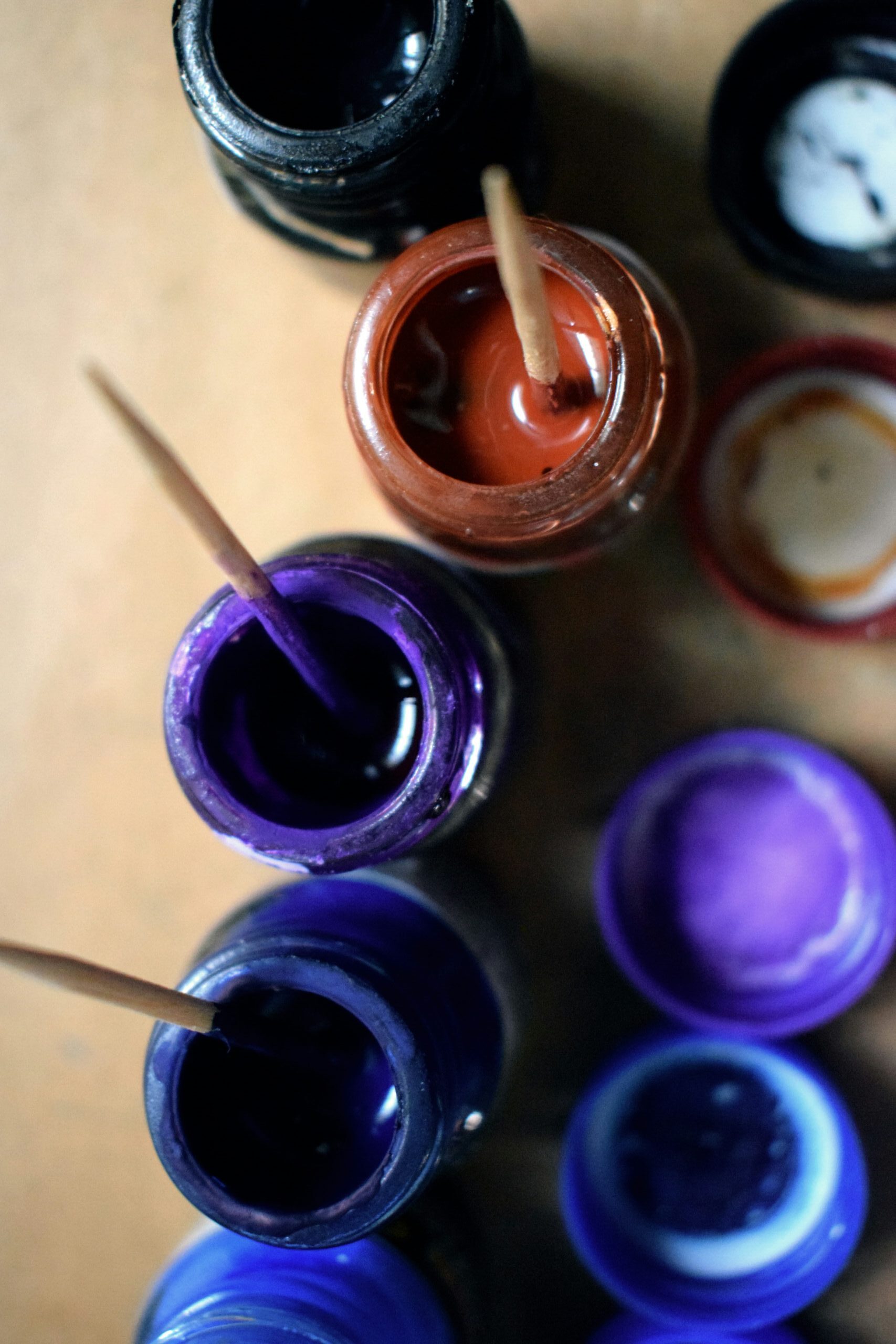 Open jars of colorful paints in shades of blue, purple, brown, and black with wooden sticks dipped inside.
